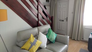 Un salon avec un canapé gris, un escalier en bois, une porte blanche et des rideaux beige.