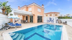 Ein Schwimmbecken mit Fliesenplatten, eine überdachte Terrasse mit Tisch und Stühlen sowie ein Gebäude mit einem gefliesen Dach und Fenstern.