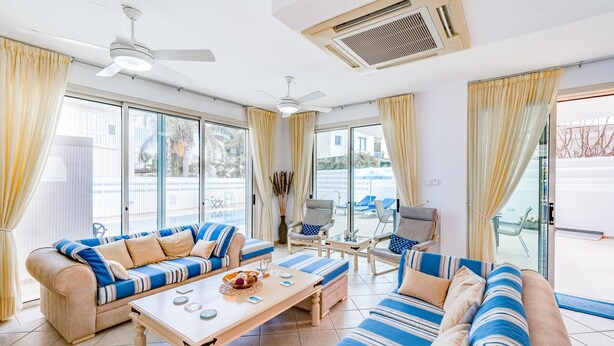Ein geräumiges Wohnzimmer mit einer Sofagarnitur, einem Couchtisch und einem Esstisch. Es gibt Deckenventilatoren, große Fenster mit Vorhängen und einen Blick ins Freie.