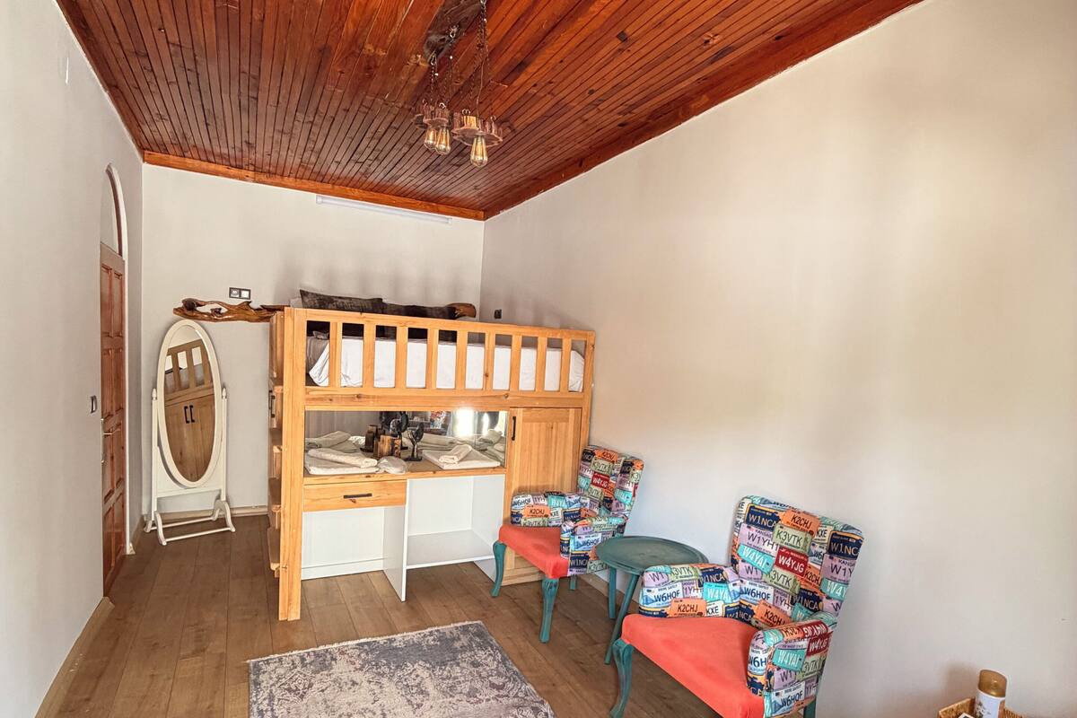Ein Zimmer mit Holzbalkendecke, einem Etagenbett, einem Schreibtisch, zwei Stühlen mit bunten Mustern, einem Teppich und einem Fernseher.