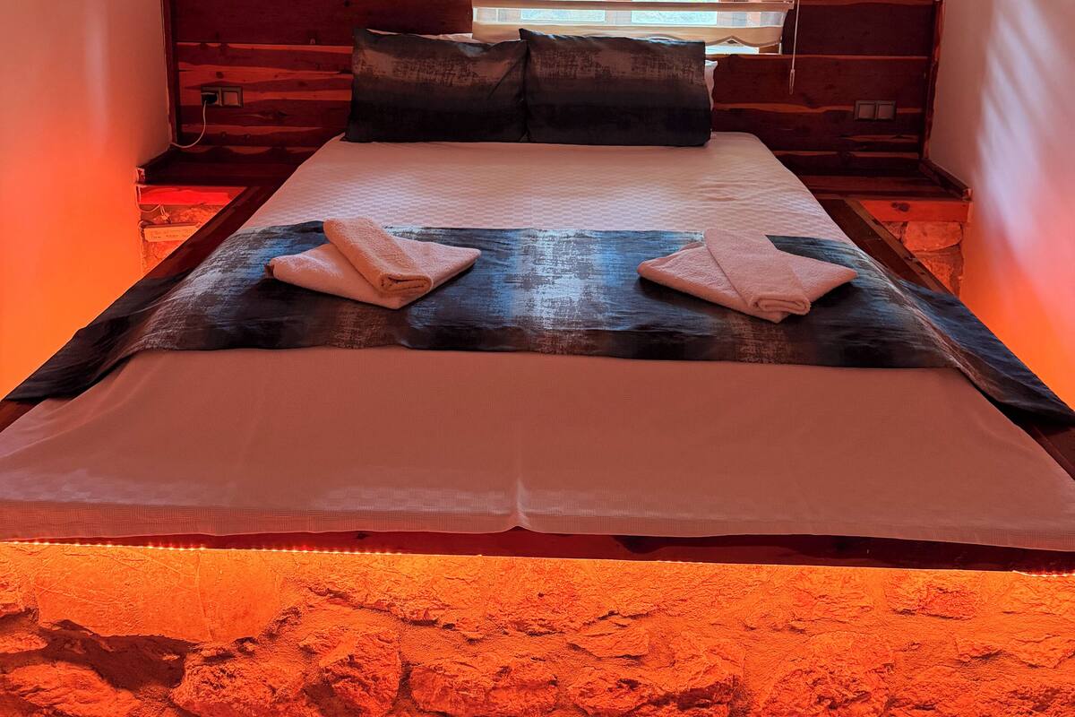 Ein Schlafzimmer mit einem großen Bett, einem Fenster mit Jalousien, einem Nachttisch und einer Klimaanlage.