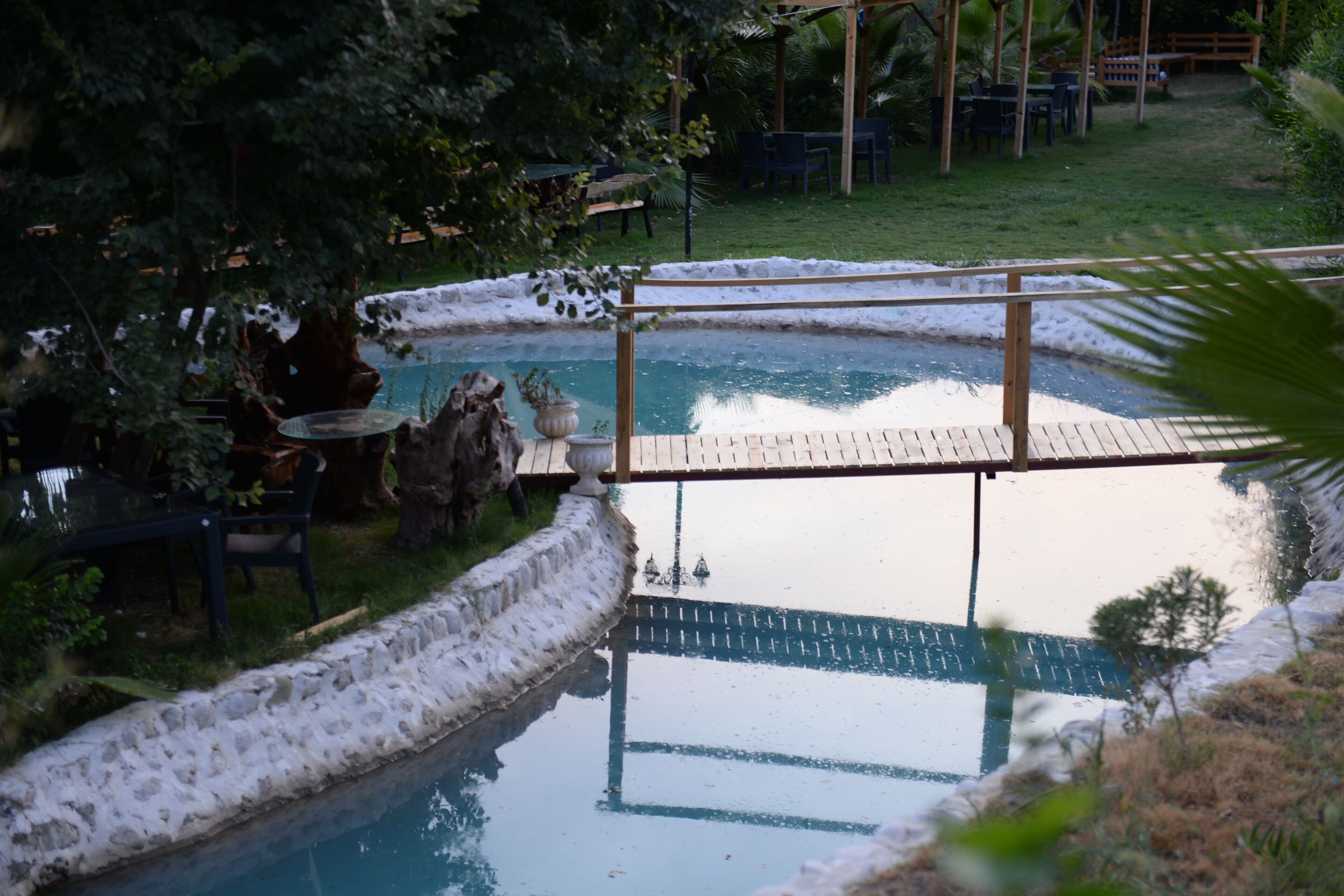 A pool with a wooden deck and stone edges, surrounded by greenery and outdoor furniture.