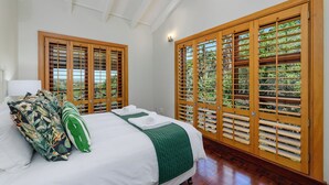 A bedroom with a bed, pillows, a green throw blanket, wooden shutters, and a view of greenery.