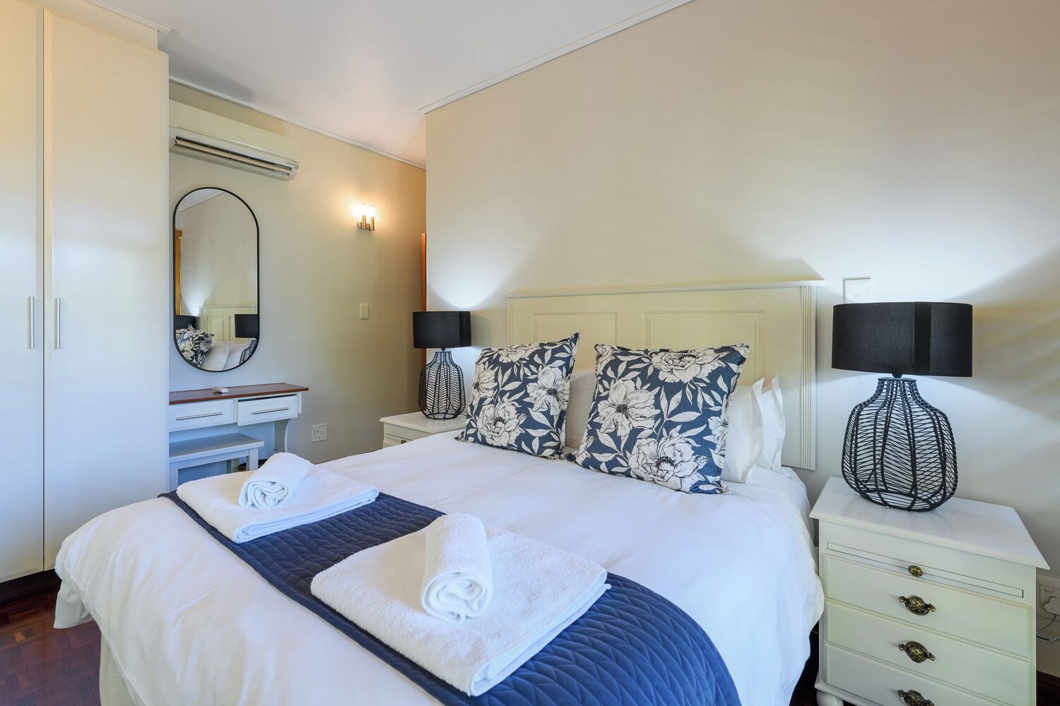 A neatly made bed with white and blue floral pillows, a bedside table with a lamp, and a mirror on the wall.