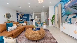 A modern living room with a brown sectional sofa, a round woven ottoman, and a large painting on the wall.