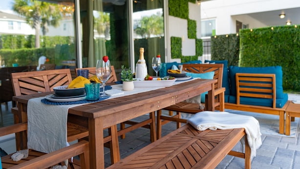 An outdoor dining area with wooden furniture, a table set with plates, glasses, and a bottle of champagne.