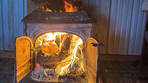 Ein Kaminofen mit Feuer, ein Holzregal mit Gegenständen darauf und eine wellenförmige Metallwand im Hintergrund.
