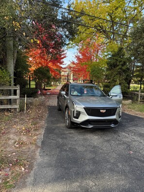 Un SUV Cadillac parcheggiato su un vialetto asfaltato con un cancello, circondato da alberi con foglie autunnali.
