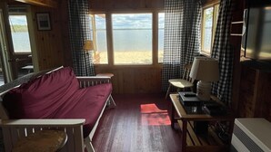 Una cabaña con interior de madera, una cama, una mesa con lámpara, un televisor y vistas a la playa.