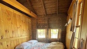 Une chambre de style cabane avec un intérieur en bois, un lit recouvert d’une courtepointe, une table de chevet et une fenêtre avec des rideaux.