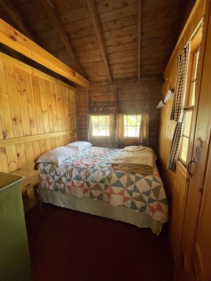 A cabin-style room with a wooden interior, a bed with a quilt, a nightstand, and a window with curtains.