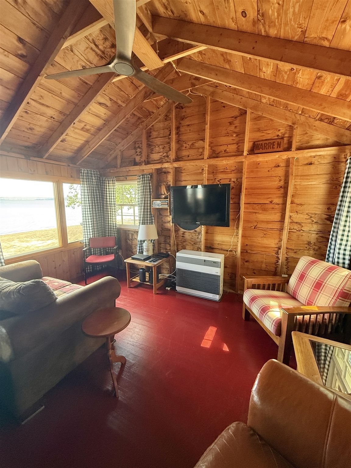 A cabin interior with a ceiling fan, a television mounted on the wall, a couch, a chair, and a small table with a lamp.