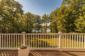 A wooden deck with a white railing, overlooking a grassy area with trees and a body of water in the background.