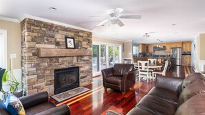 A spacious living room with a stone fireplace, leather sofas, and a dining area with a kitchen in the background. - WATERFRONT ON LONG LAKE  (HARRISON)