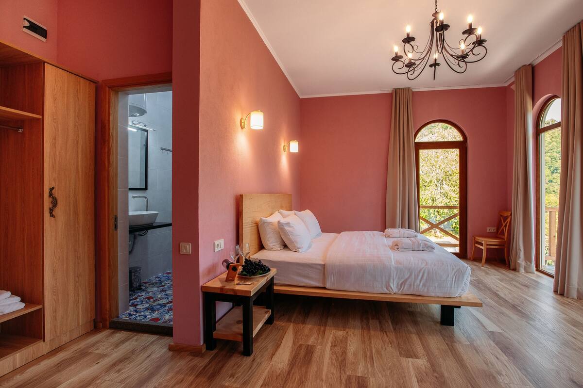 Une chambre avec un lit en bois, une table de chevet sur laquelle se trouve un panier de fruits, une commode et une vue sur la salle de bain par une porte ouverte.