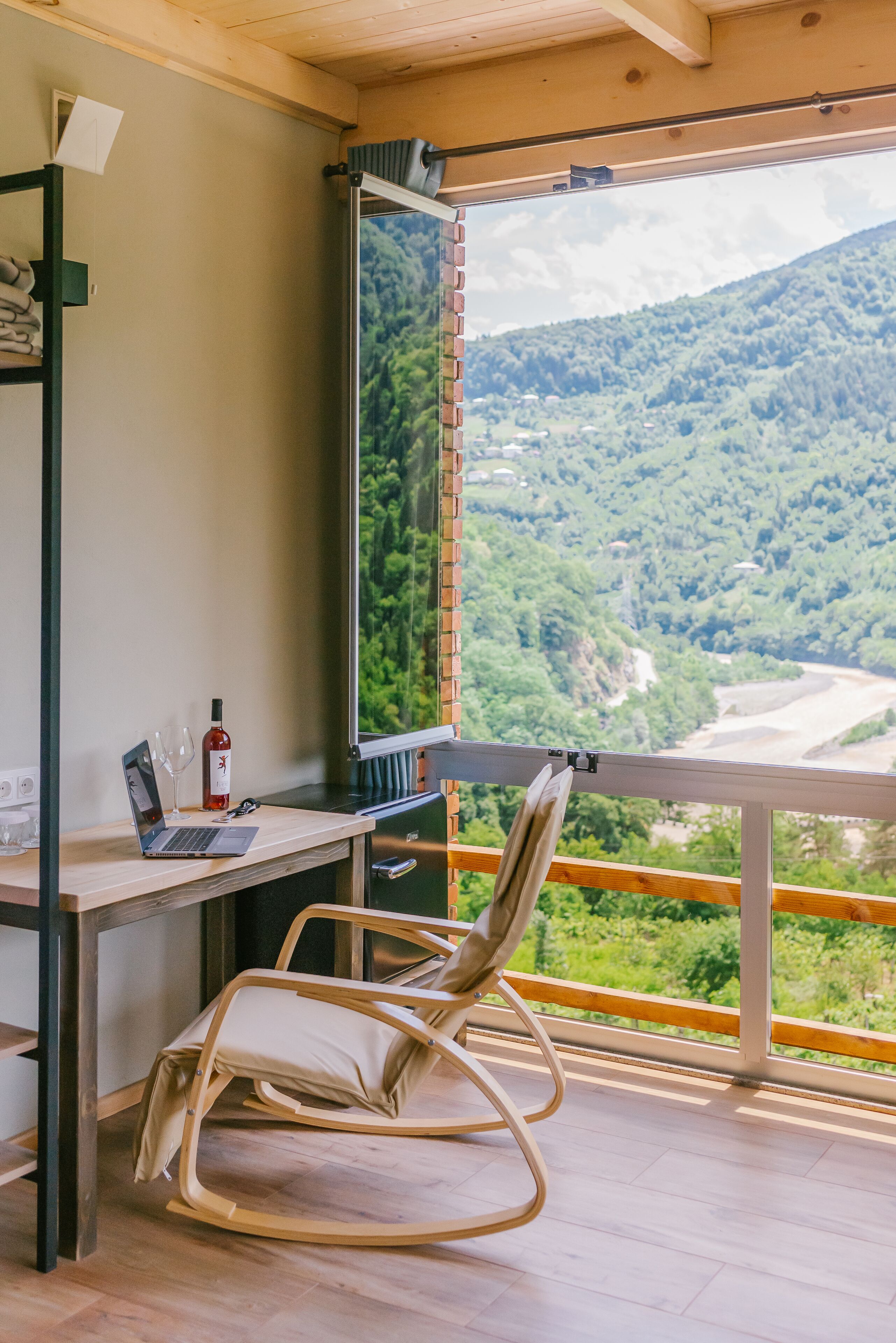 A room with a desk, laptop, wine bottle, and glasses, with a view of a forested valley through a large window.