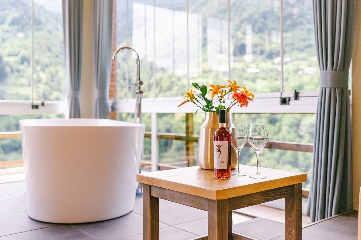Une salle de bain avec une baignoire blanche, une table en bois sur laquelle se trouvent une bouteille et des verres, et une vue sur la verdure à travers de grandes fenêtres.