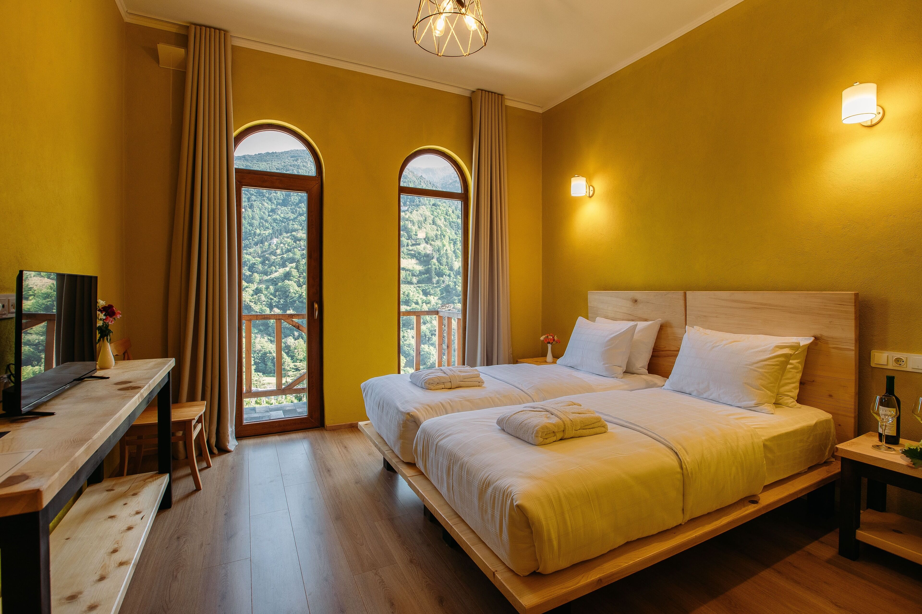 A hotel room with a large bed, a desk with a television, and a view of the outdoors through arched windows.