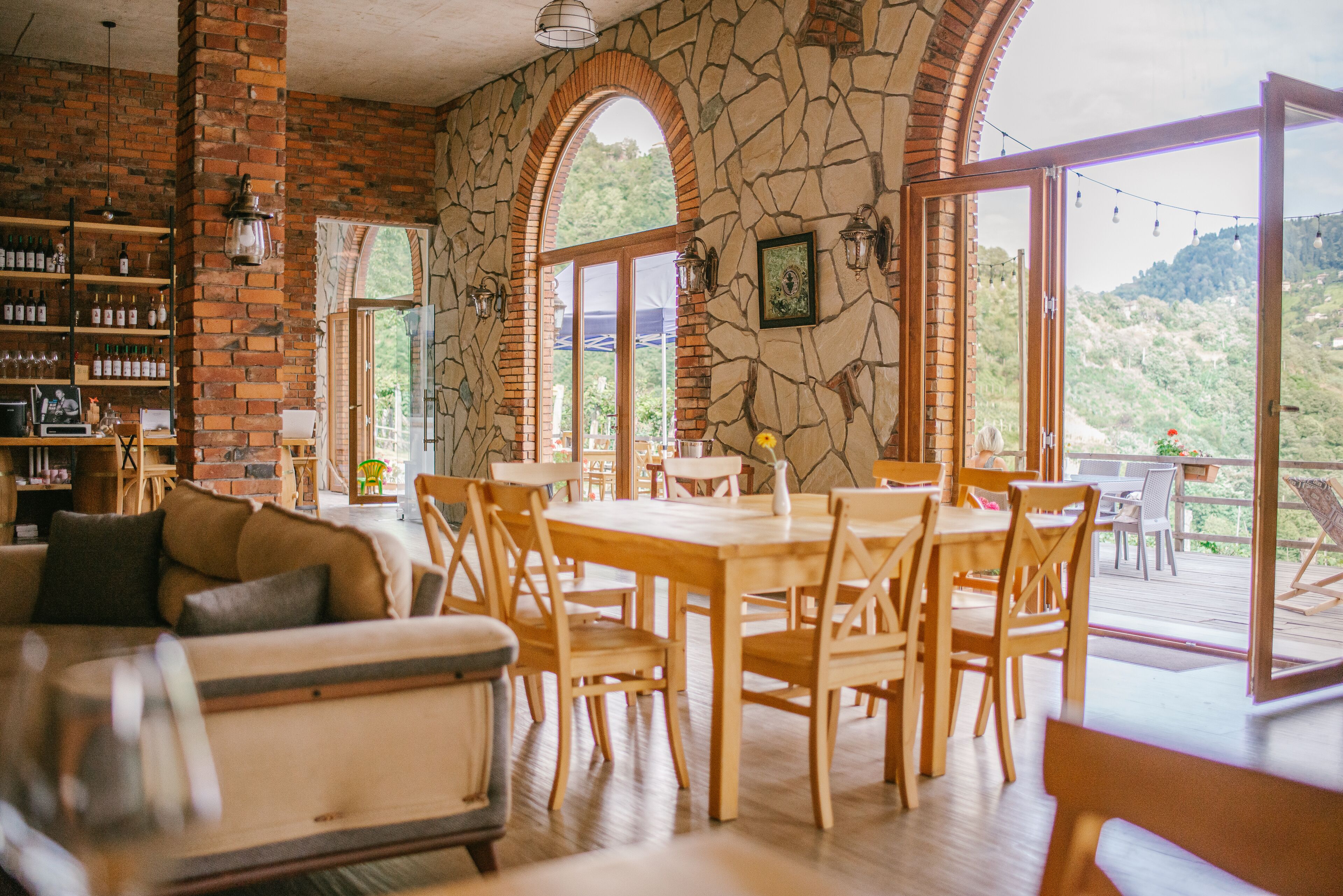 Un comedor con pared de piedra, mobiliario de madera y grandes ventanales con vistas a un paisaje pintoresco.