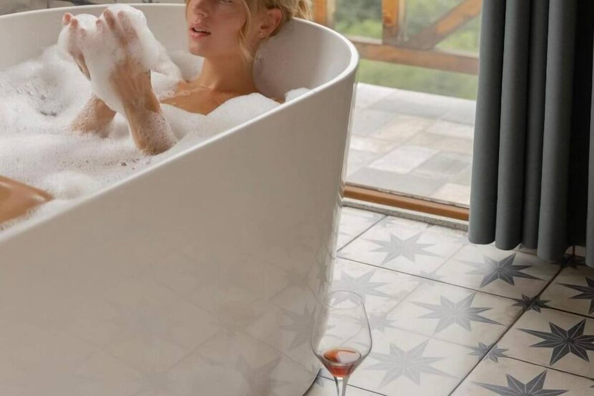 Une personne se détend dans une baignoire, le sol étant décoré de motifs en forme d’étoiles, et on aperçoit une terrasse en bois à travers la fenêtre.