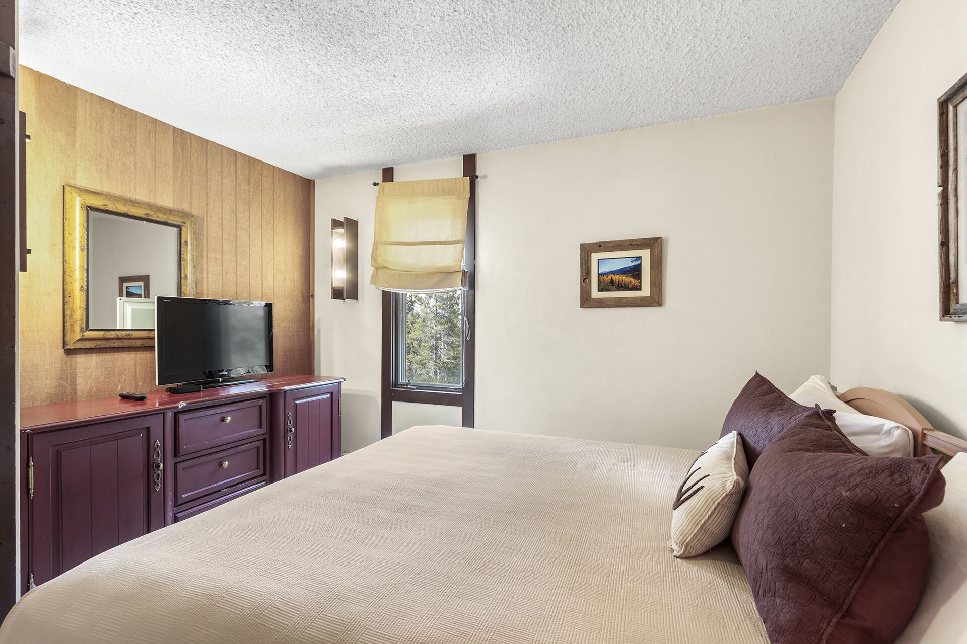 A hotel room with a bed, a TV on a wooden dresser, a window with curtains, and a framed picture on the wall.