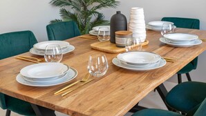 Une table à manger dressée avec de l’assiette blanche, des verres transparents et des couverts dorés, entourée de chaises vertes.