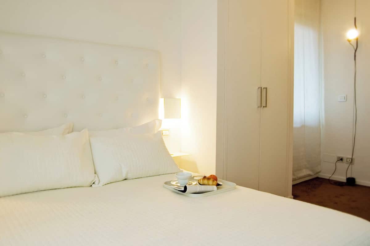 A hotel room with a large bed, a bedside table with a lamp, and a tray with a croissant and a cup.