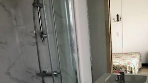 Ein Badezimmer mit Duschkabine aus Glas, Waschbecken mit Spiegel und einem Bett mit floralem Überwurf im Hintergrund.