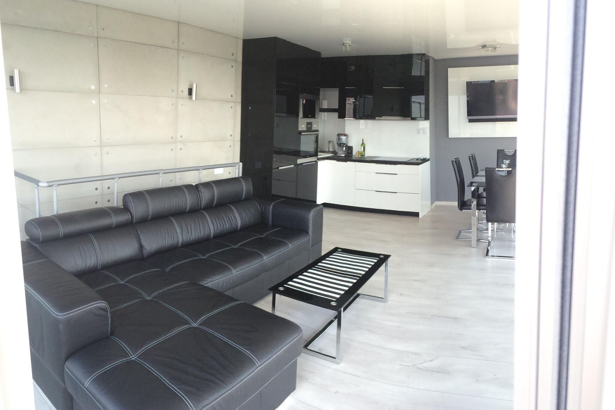Un salon moderne avec un canapé en cuir noir, une table basse en métal et un coin cuisine doté de meubles blancs et d’un plan de travail noir.