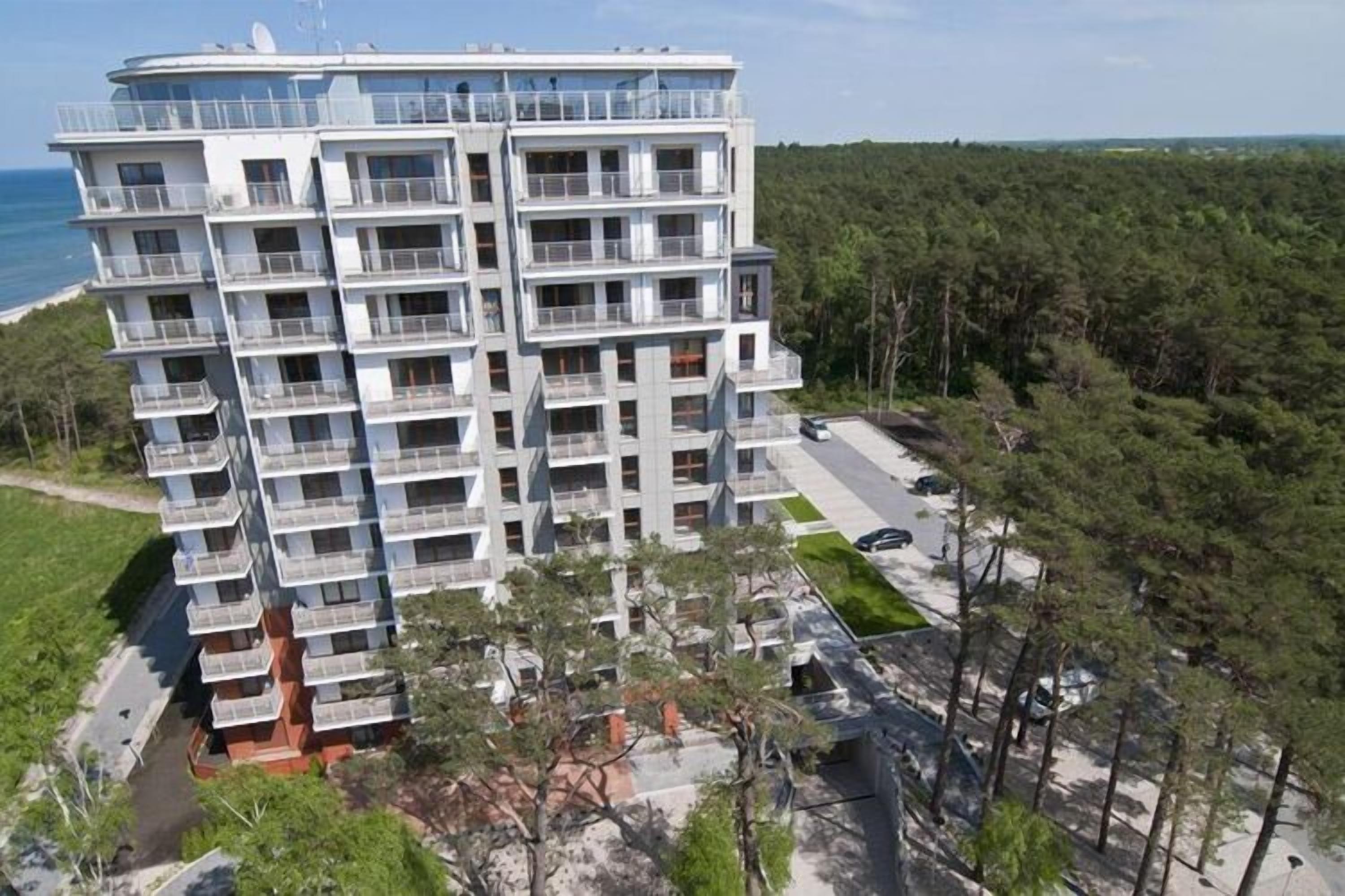 A multi-story residential building with balconies, located near a forested area and a body of water.