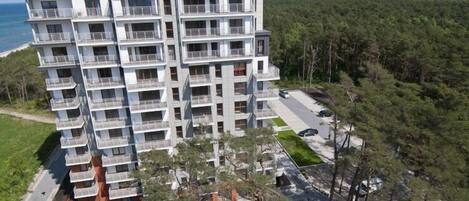 A multi-story residential building with balconies, located near a forested area and a body of water.