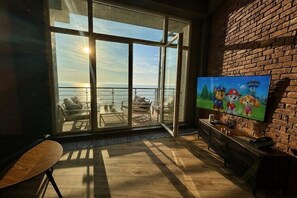 A room with a large window overlooking the sea, a television, and a wooden floor.