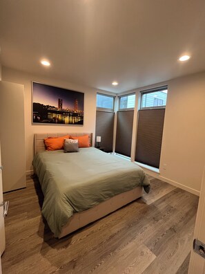 Un dormitorio con cama, un cuadro de paisaje urbano, una ventana con persianas y una puerta.
