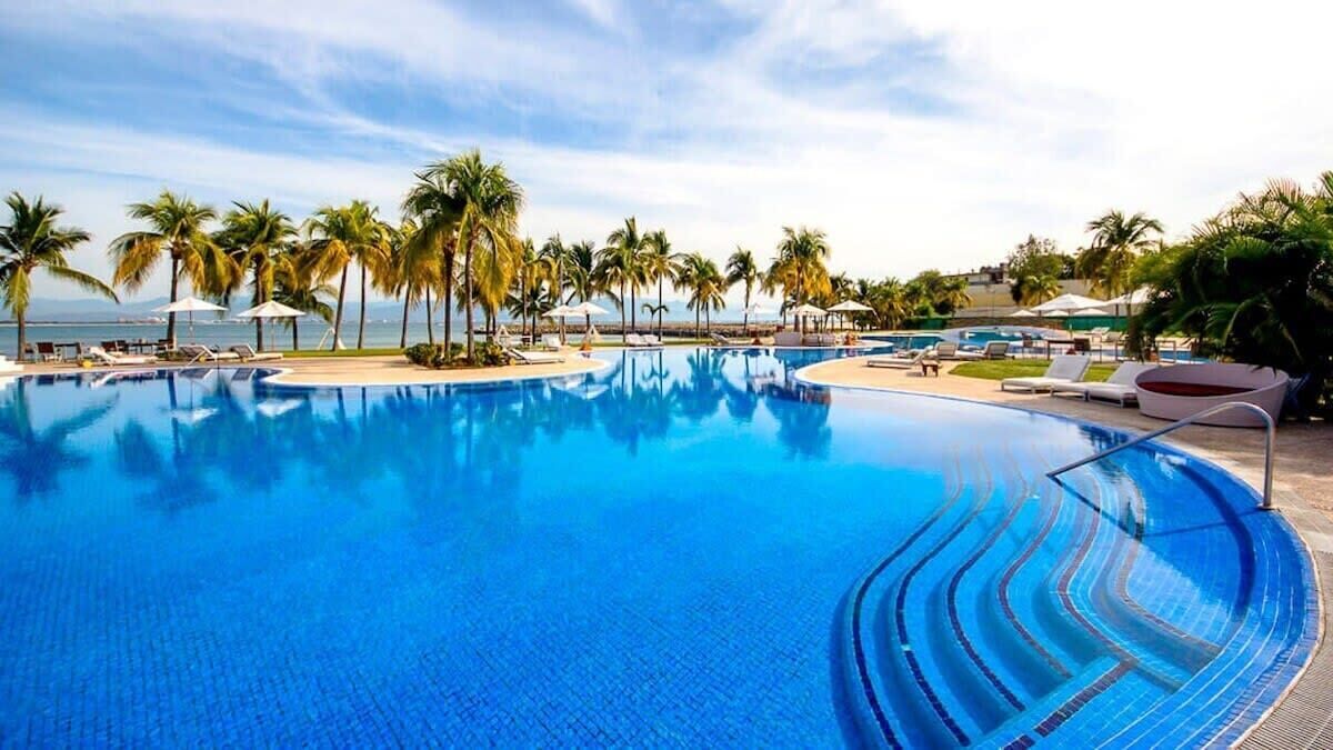 Una piscina grande y cristalina de color azul, rodeada de palmeras y sillas de descanso, con vista al océano al fondo.