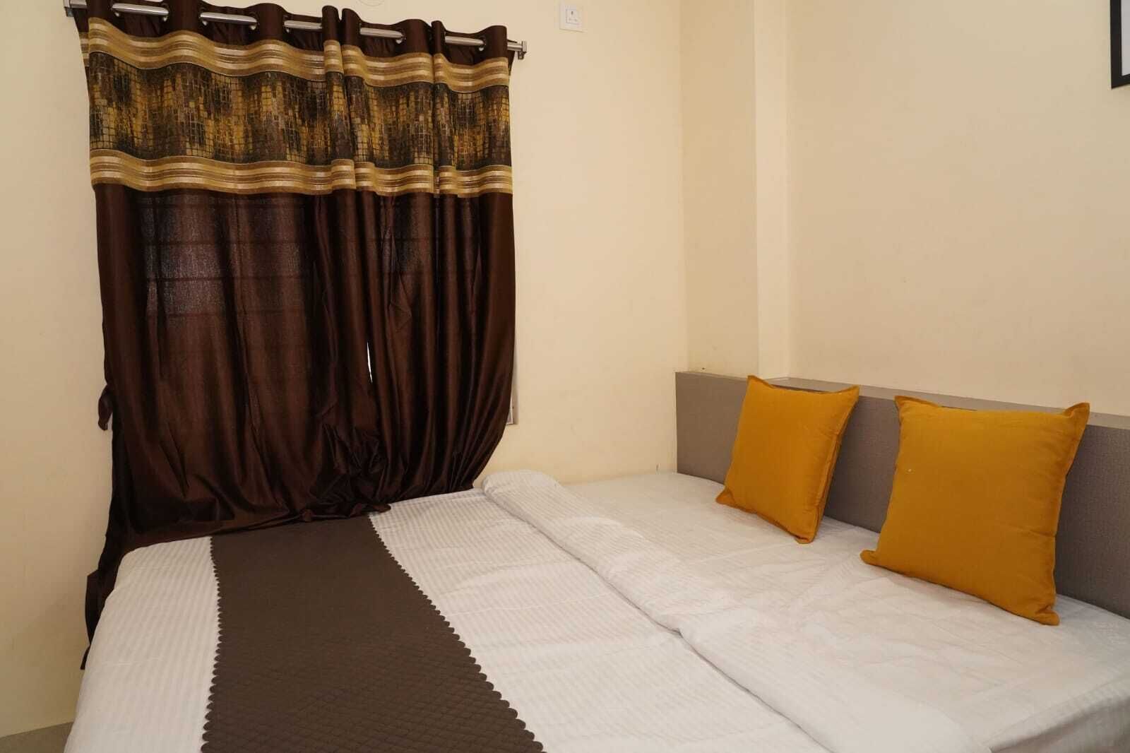 Uma cama de solteiro com uma colcha branca e duas almofadas amarelas mostarda, uma cortina marrom escura com padrão dourado, e uma parede lisa com um interruptor de luz.