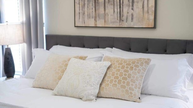 A neatly made bed with white and patterned pillows, a grey upholstered headboard, and a painting on the wall.