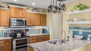 A kitchen with wooden cabinets, a stainless steel microwave, a double oven, a granite countertop, a sink, and hanging pots.