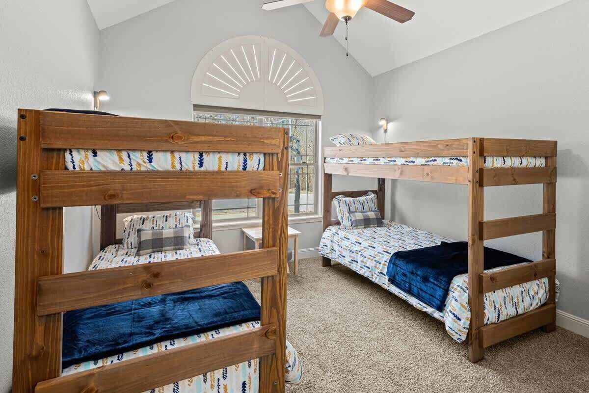 A room with two bunk beds, a ceiling fan, a window with a sunburst design, and a small table.