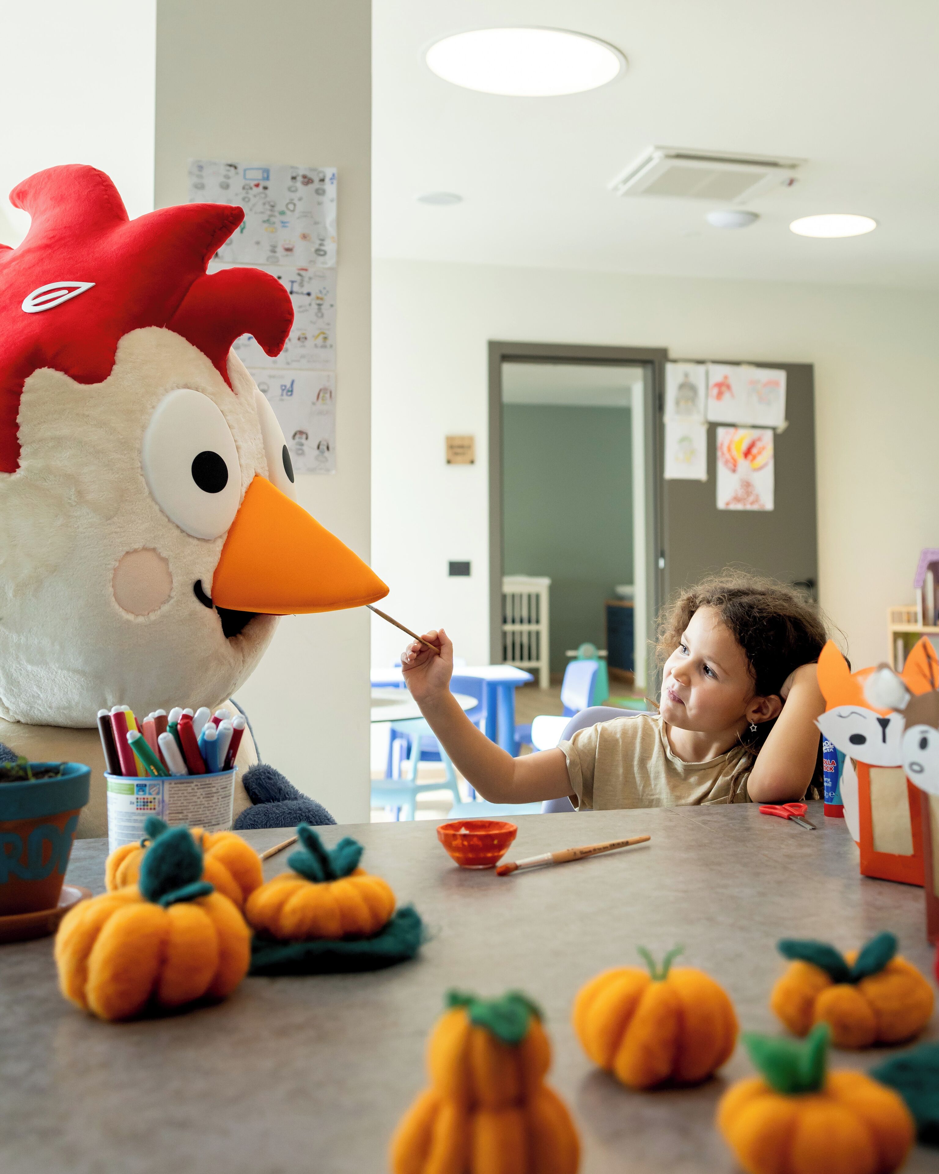 Un bambino interagisce con un grande pupazzo a forma di gallina in una stanza con un tavolo, zucche e diversi oggetti.