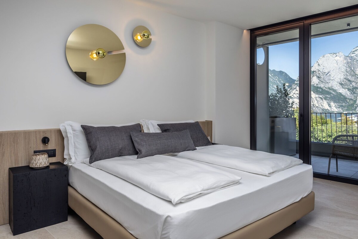 A modern bedroom with a large bed, two bedside tables, a round mirror, and a view of mountains through a sliding glass door.