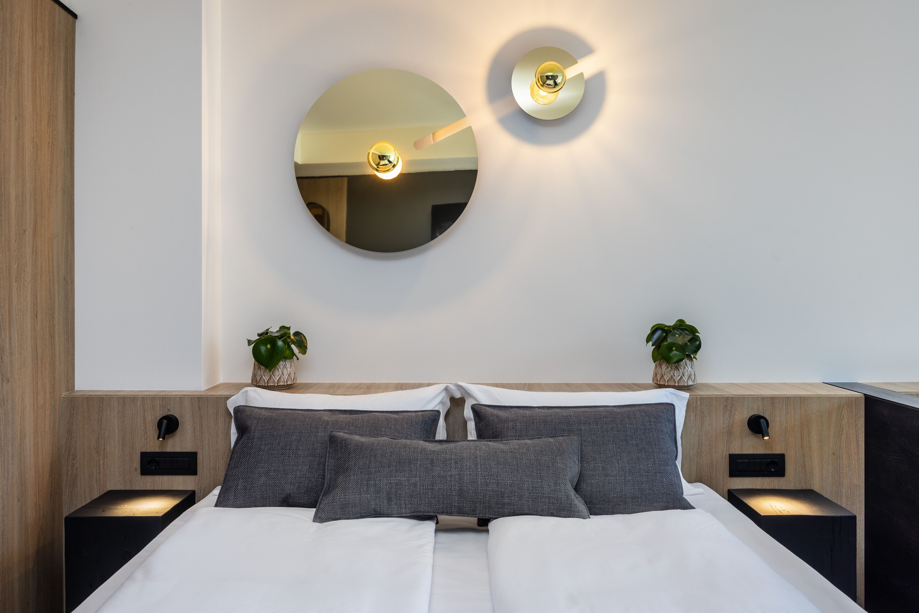 A modern bedroom with a large bed, two bedside tables with lamps, a round mirror, and two potted plants.