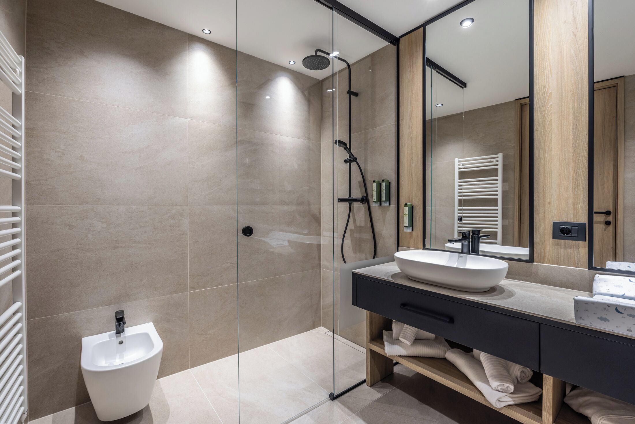 Un baño moderno con ducha de vidrio, un lavatório pedestal blanco, un espejo grande y un tocador de madera con lavabo.