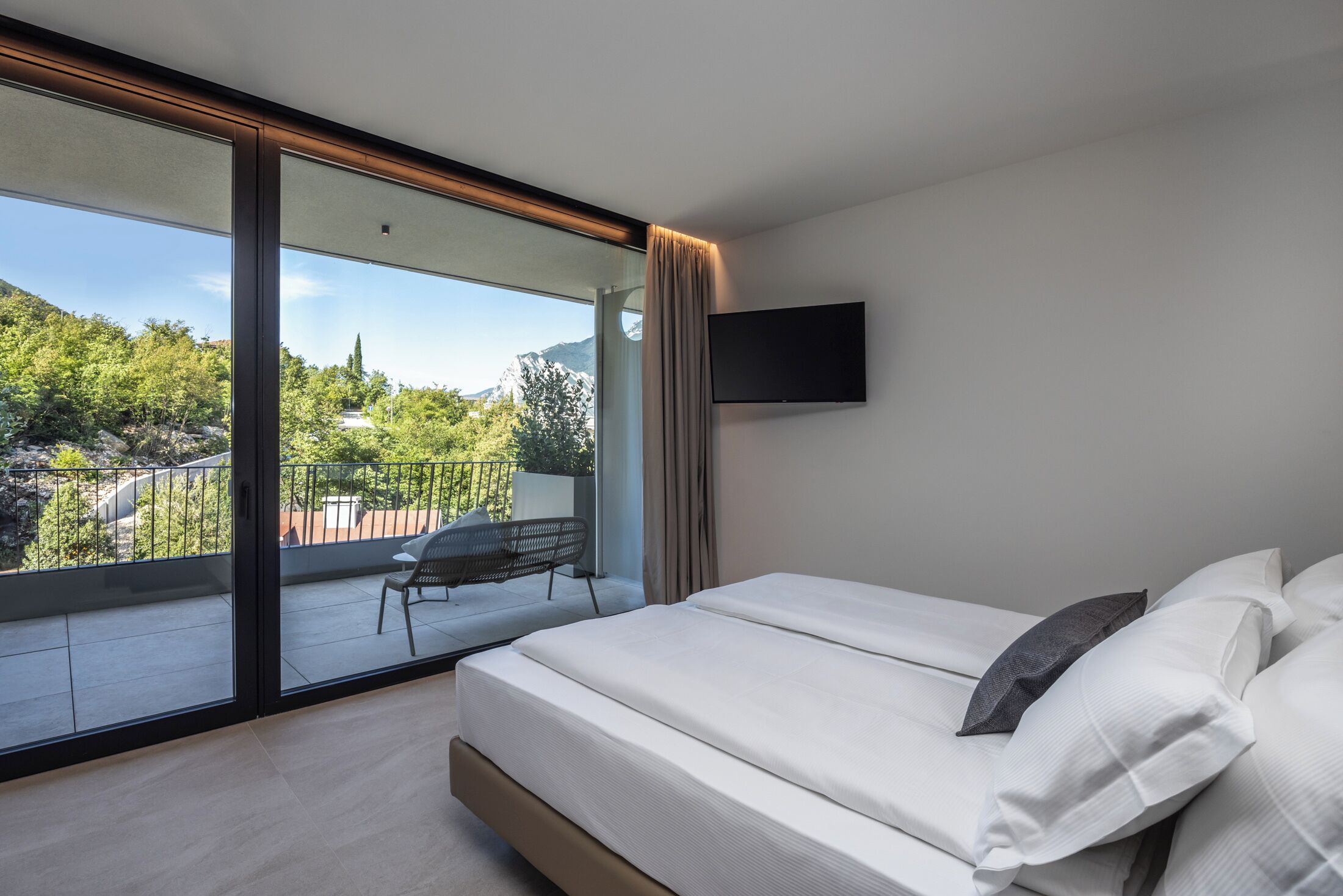 A bedroom with a large sliding glass door leading to a balcony, a bed with white linens, and a mounted TV on the wall.