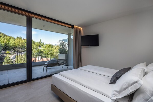 A bedroom with a large sliding glass door leading to a balcony, a bed with white linens, and a mounted TV on the wall. - Gardea Soulfamily Resort (Nago-Torbole)