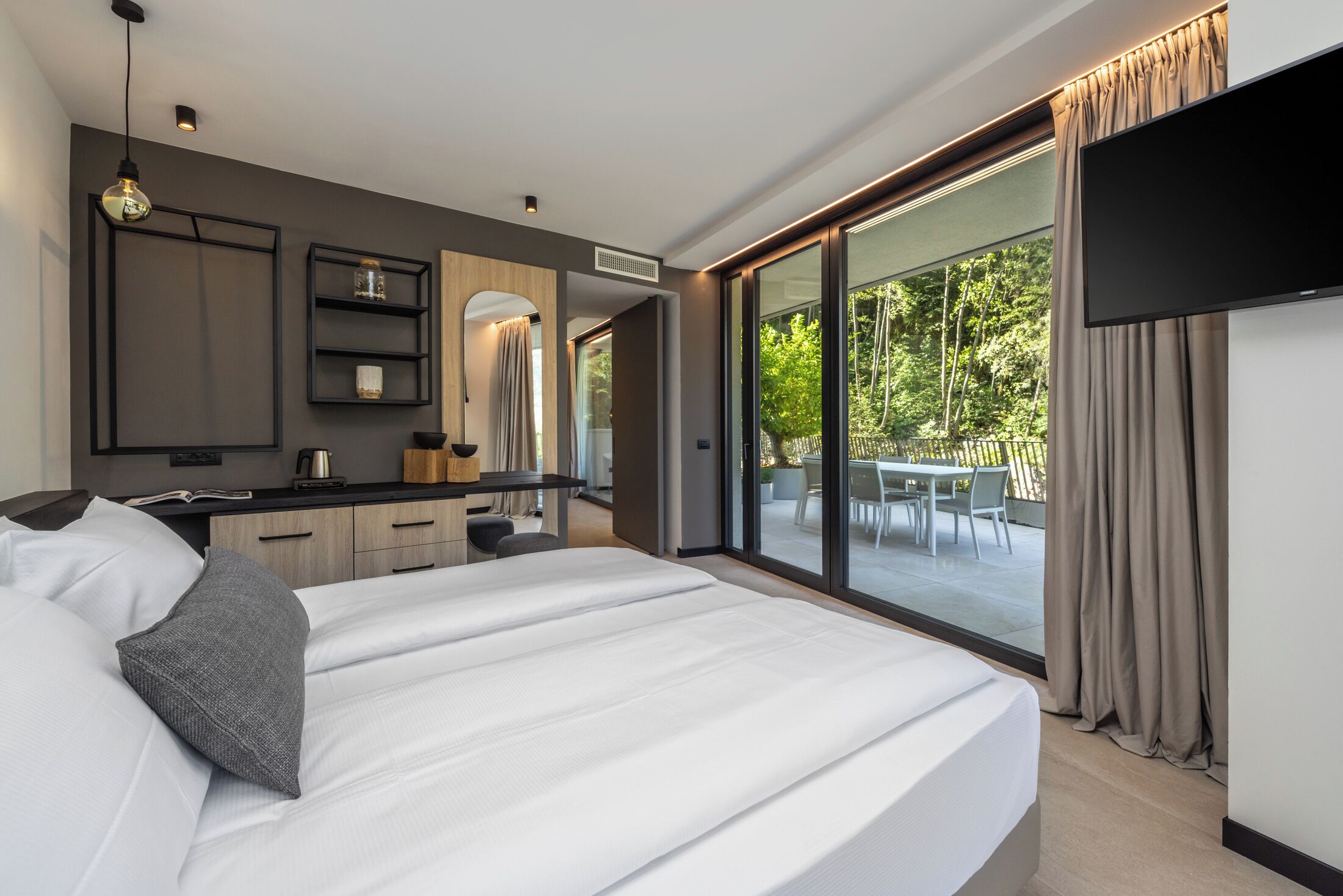 Ein modernes Hotelzimmer mit einem großen Bett, einem Flachbildfernseher, einem Essbereich mit Tisch und Stühlen sowie einer Glasschiebetür, die auf eine Terrasse im Freien führt.