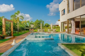 A swimming pool with a wooden deck and lounge chairs, surrounded by greenery and a modern building.