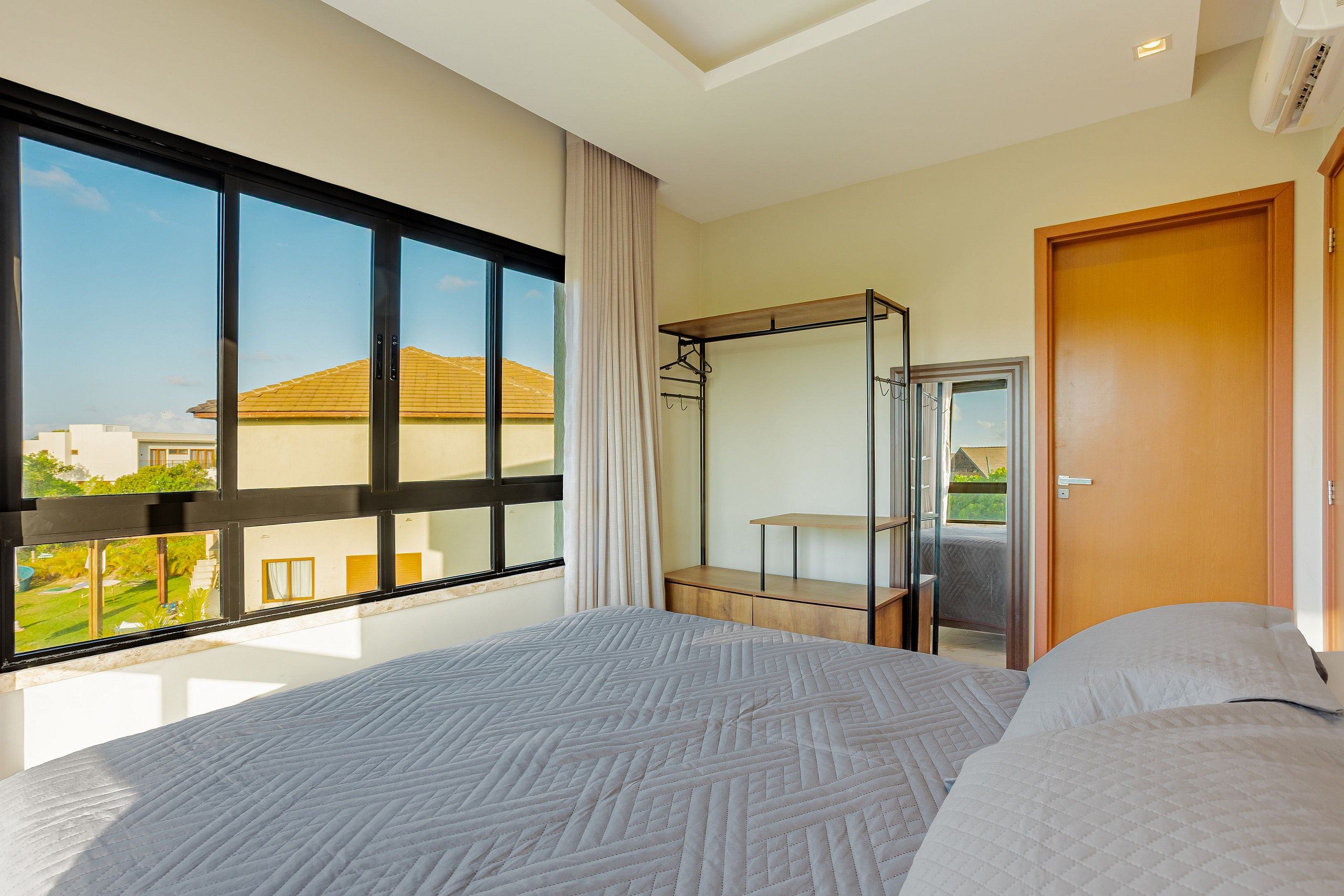 A hotel room with a large bed, a wardrobe, a mirror, and a window with a view of buildings and greenery.