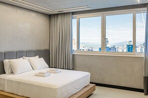 Quarto de hotel moderno com uma cama grande, cortinas cinzas e vista para a cidade pela janela.