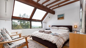 Ein Schlafzimmer mit einem großen Oberlicht, einem Bett mit karierter Bettdecke, einem Holzstuhl und einem Nachttisch mit Lampe.