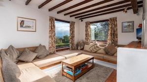 Ein gemütliches Wohnzimmer mit einer Sofagarnitur, einem Holztisch und großen Fenstern mit Vorhängen.
